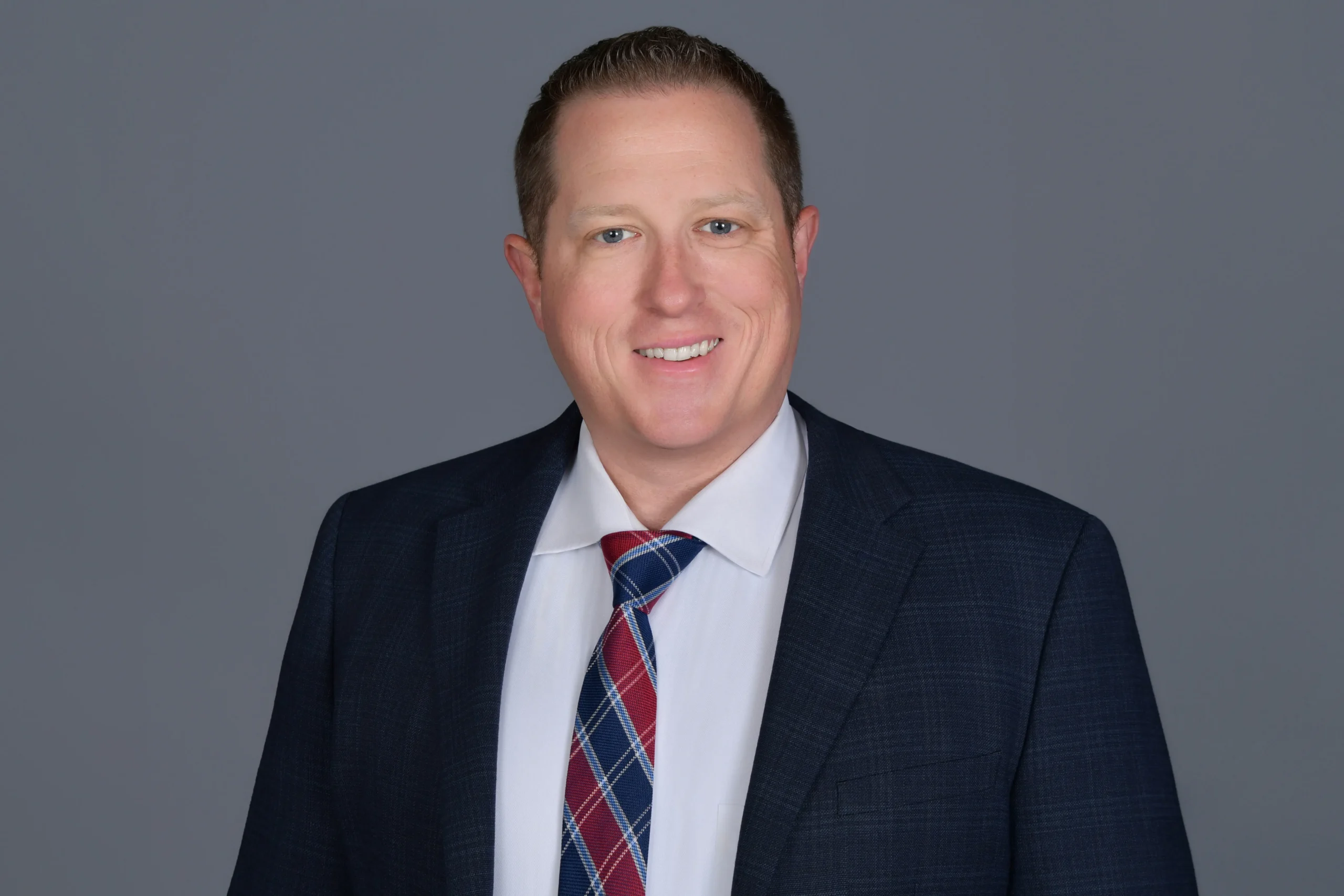 Professional portrait of a smiling man in a dark suit, white shirt, and red-blue plaid tie.