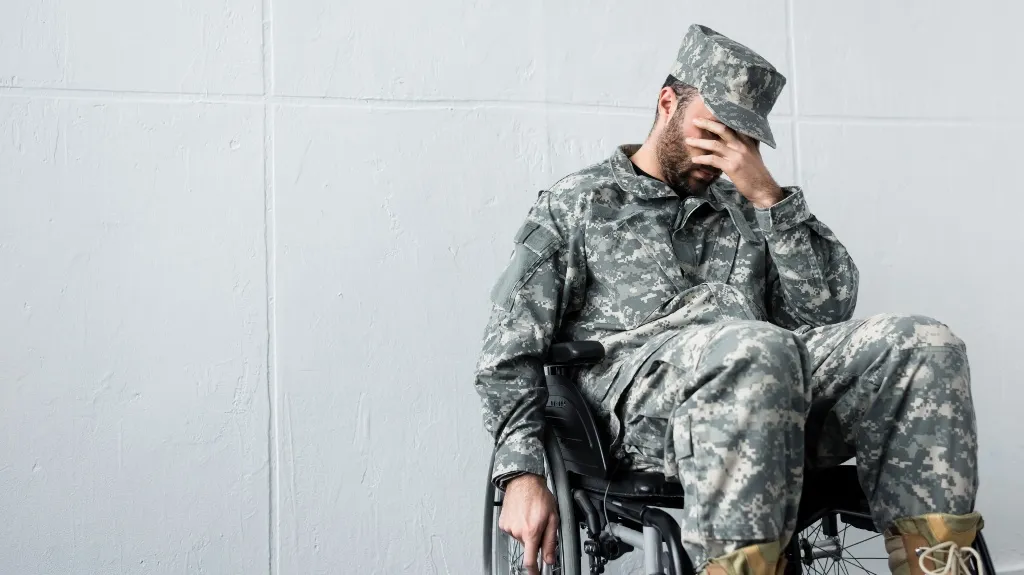 Man in digital camouflage military uniform sitting in wheelchair, covering face with hand, against white wall.