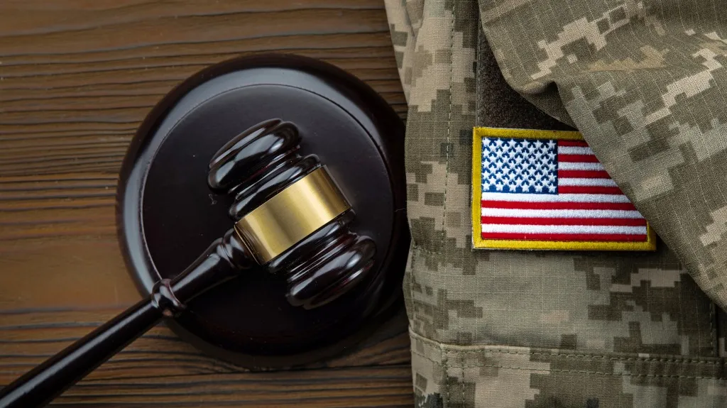 Dark wooden judge's gavel next to a U.S. flag patch on camouflage military uniform.